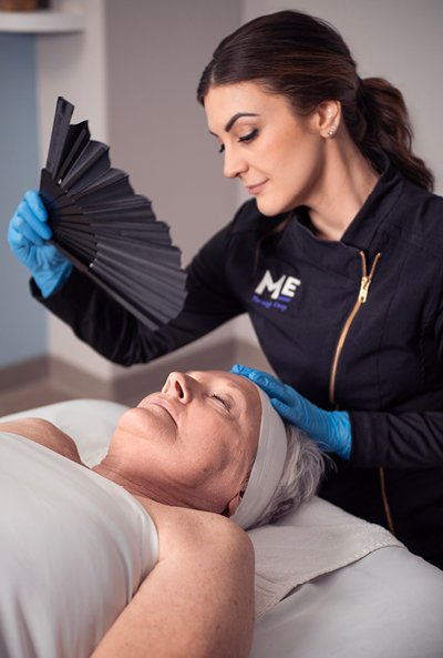 Woman laying on table as esthetician waves fan above her face. 