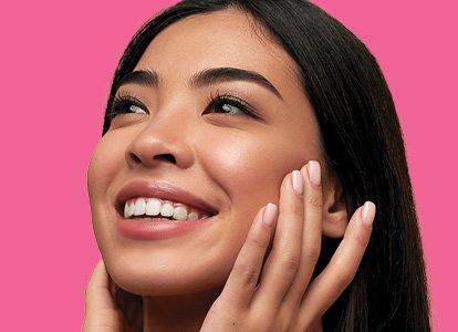 Close-up of woman touching her face with both hands in front of a pink background. 