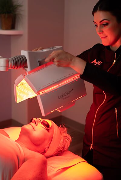 Esthetician standing above a woman lying on table and receiving an Age-Defying Nourishing Light Treatment. 