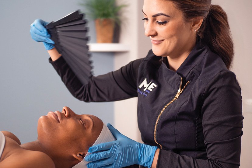 Esthetician smiling as she waves a fan over a client during a hydrafacial treatment. 